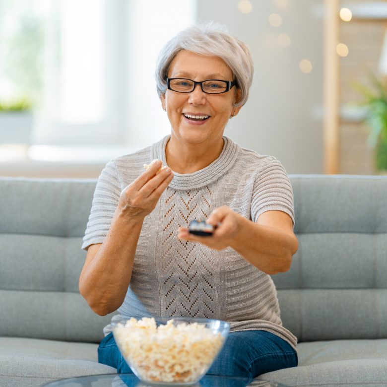 Senior woman sitting on a couch, holding a remote control and eating popcorn