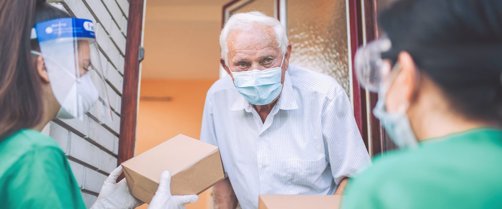 J2523_Face_Mask_1680x700 An elderly man in a mask receives packages from two healthcare workers wearing face shields and gloves at his doorstep.