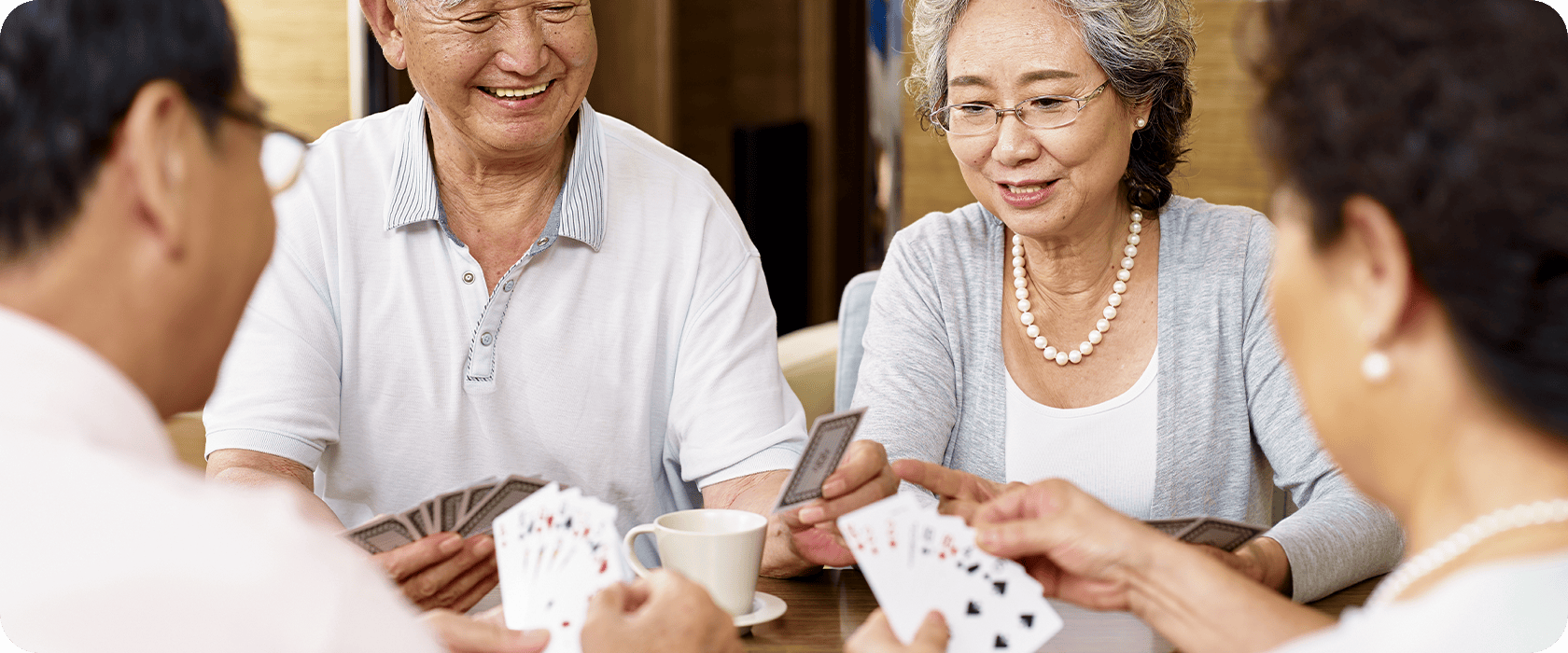 Four people play cards at a table, smiling and interacting. A cup of coffee sits on the table, indicating a casual, friendly gathering.