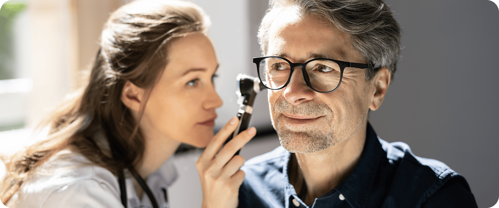 J3607_Header 1 A doctor examines a man's ear using an otoscope in a well-lit room. The man is wearing glasses and appears relaxed while the doctor focuses intently.