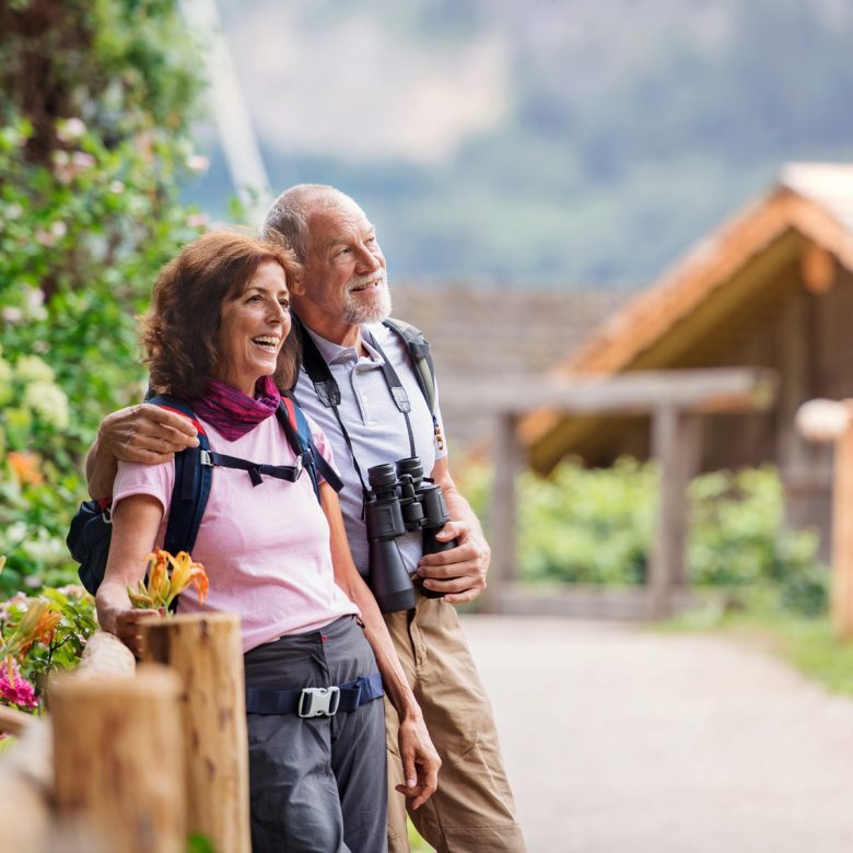J2625_SummerTravel_1200x1200-780×780 Senior man and woman strolling through a mountainous area, smiling and appreciating the natural beauty around them.