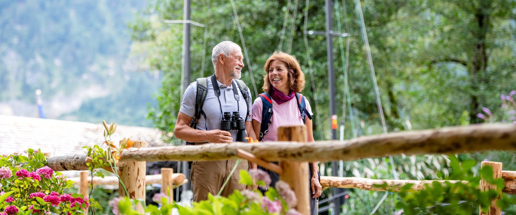 J2625_SummerTravel_1680x700 A smiling couple walks along a rustic wooden fence, holding binoculars, amidst vibrant flowers and lush greenery in a serene, natural setting.