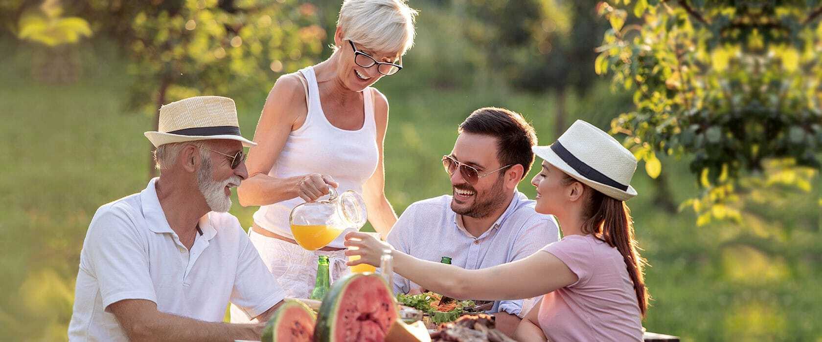 A woman pours orange juice into a glass while three people joyfully converse around an outdoor table laden with food, in a sunny garden setting.
