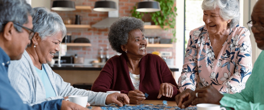 A group of five elderly individuals joyfully collaborates on assembling a puzzle at a table, engaging in lively conversation within a cozy home setting.