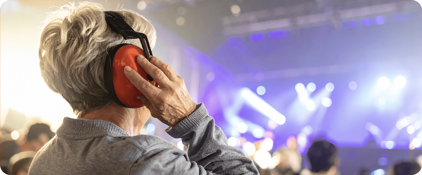 A person wearing orange earmuffs is holding them to their head while standing in a crowded, brightly lit concert venue.