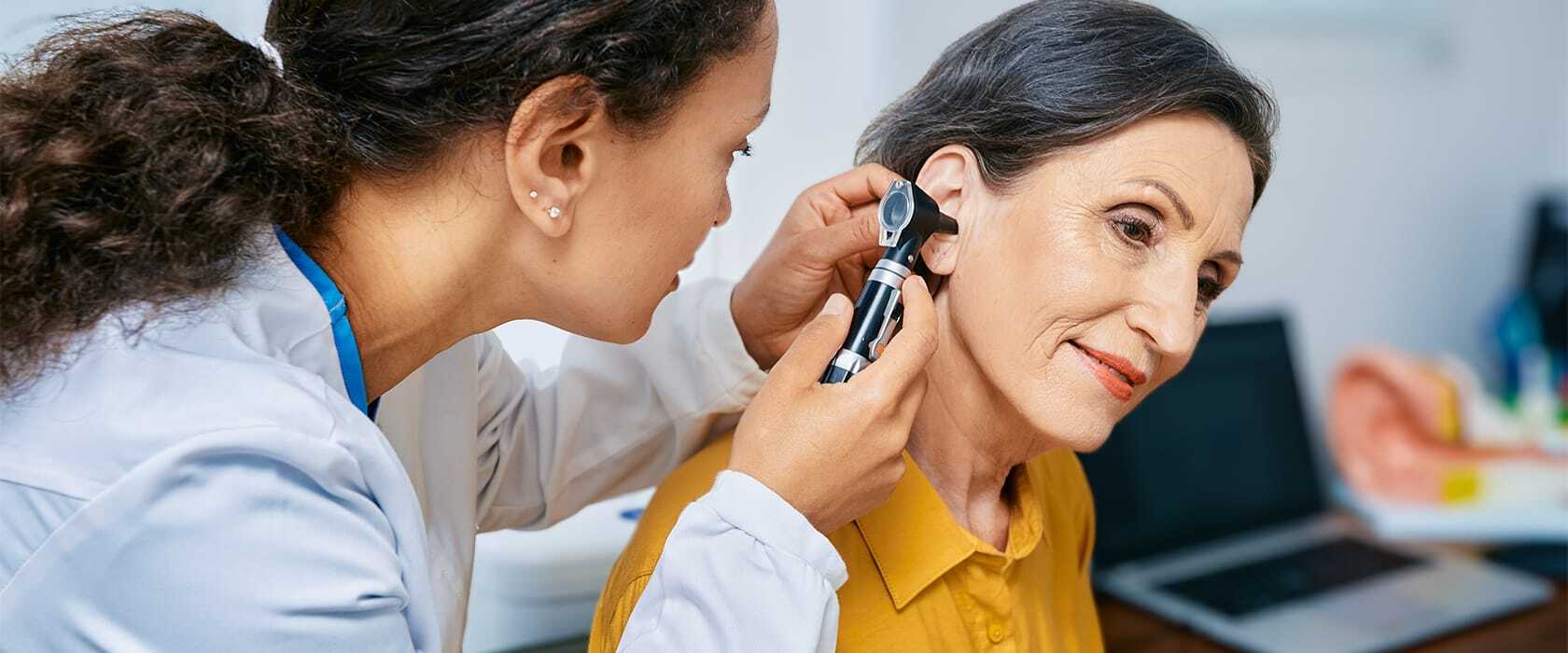 A healthcare professional is examining a patient's ear using an otoscope in a clinical setting. The patient sits calmly during the examination.