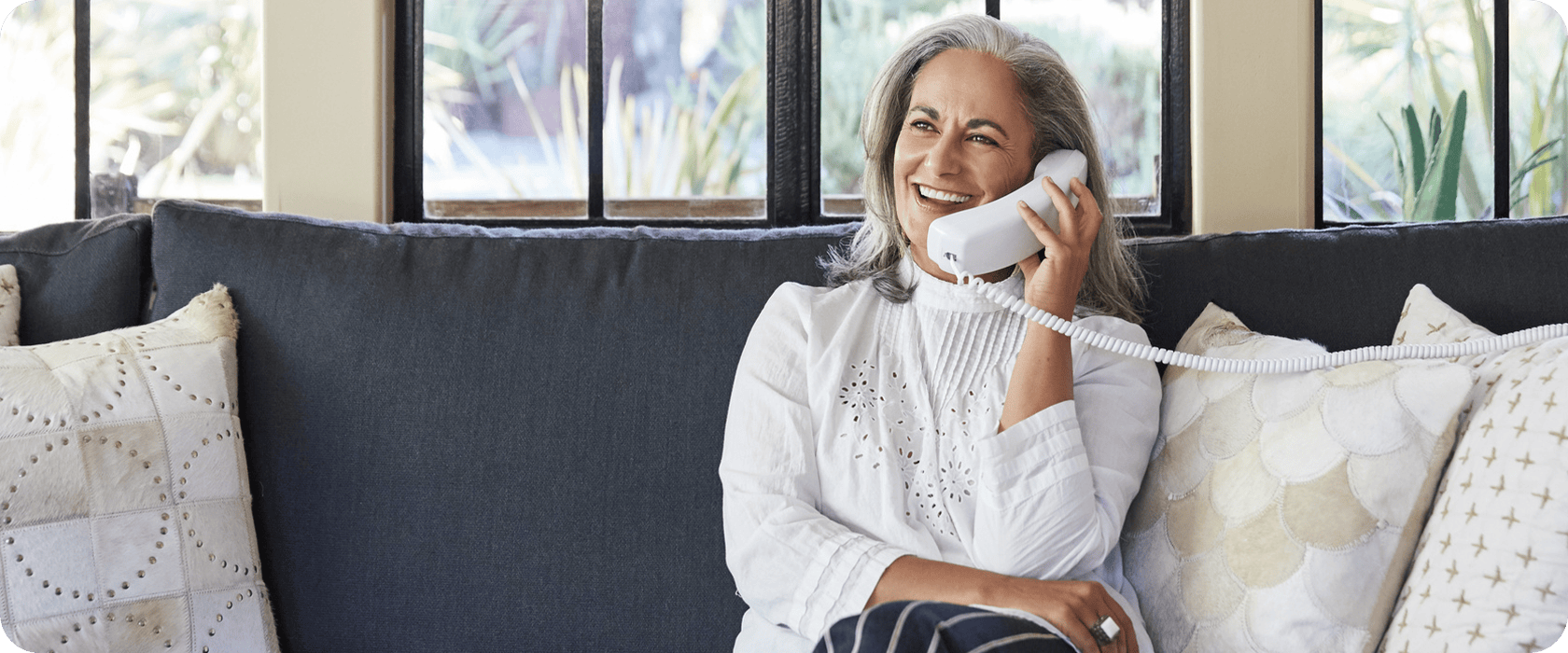 A woman with gray hair is sitting on a sofa, smiling while talking on a corded telephone amidst patterned cushions.