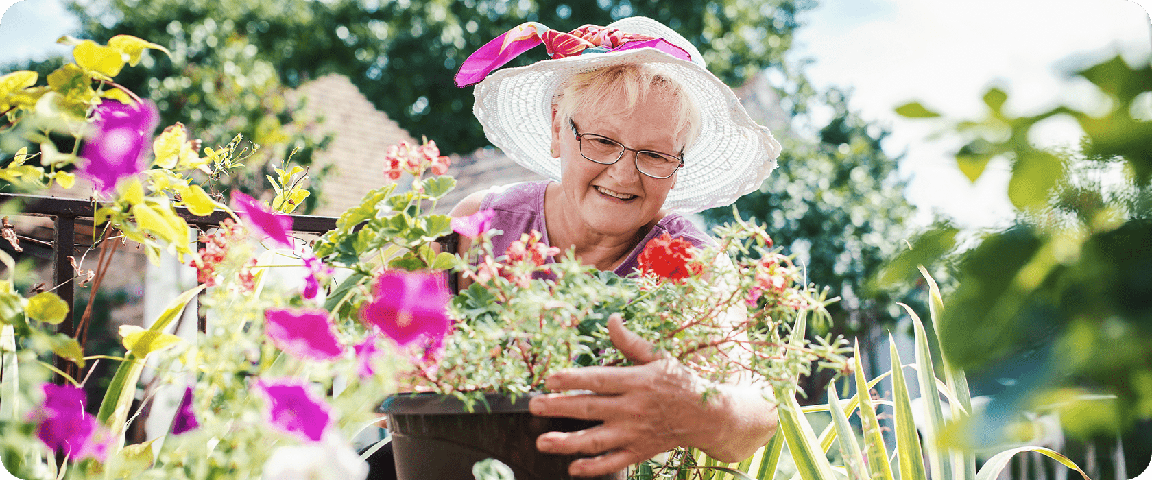 A woman in a white sunhat and glasses tends to a pot of colorful flowers outdoors, with sunlight filtering through greenery.