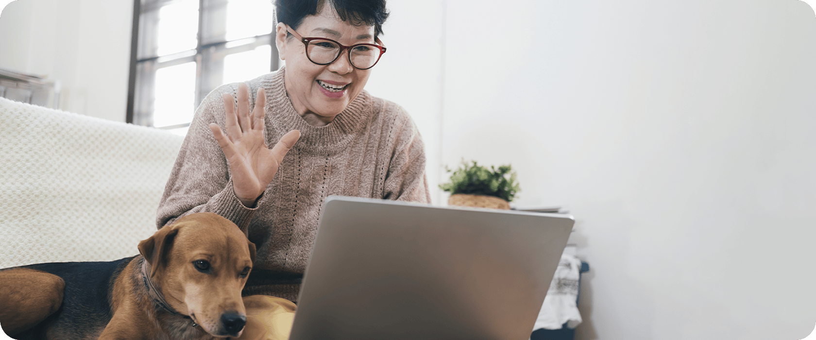 A person wearing glasses is smiling and waving at a laptop while sitting on a couch with a dog resting beside them.